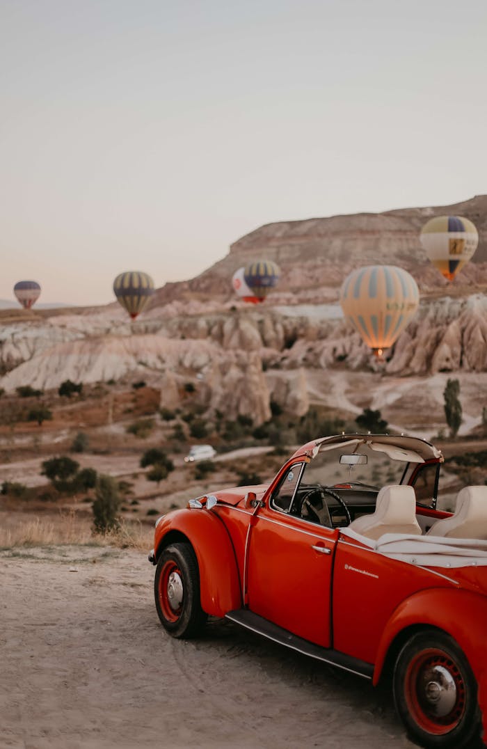 A classic red convertible parked in front of hot air balloons in Cappadocia's unique landscape.